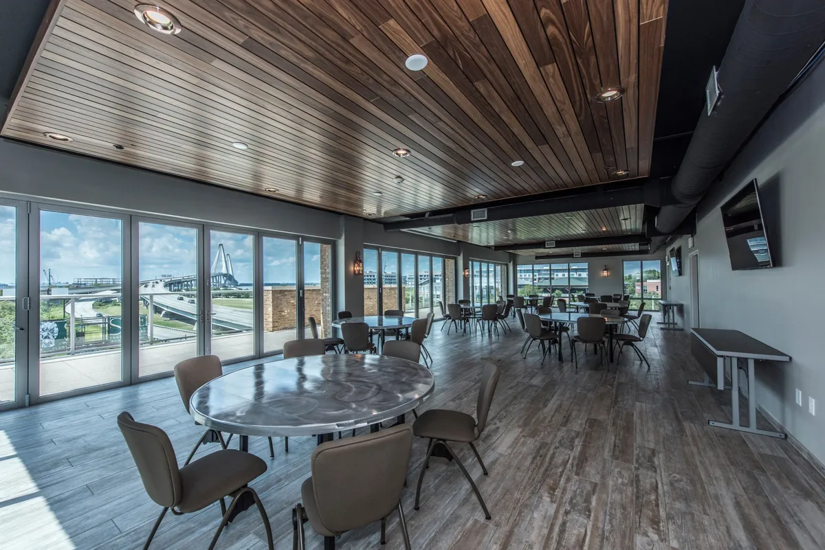 This image presents a spacious interior dining area with circular tables set beneath a stylish wooden ceiling. Large windows provide abundant natural light and offer views of the outdoor area, establishing a social and inviting atmosphere ideal for shared meals and gatherings.