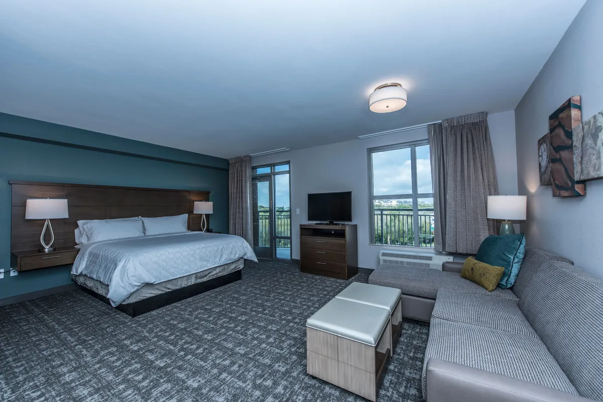This image showcases a spacious studio with a king-sized bed, a sofa seating area, a large window providing natural light and scenic views, and a television. The layout highlights comfort, relaxation, and the autonomy of a private rental.