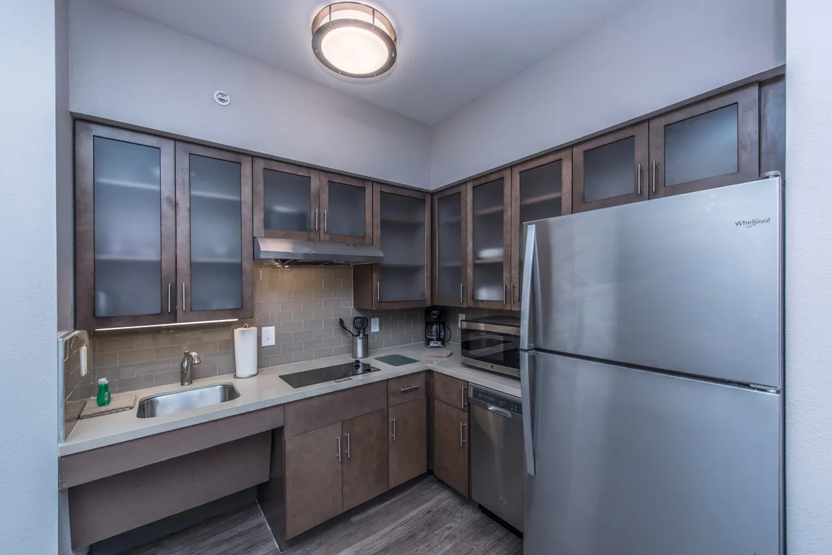 This image presents a bright, inviting kitchen featuring a stainless steel refrigerator, wooden cabinets, a well-lit countertop, and a visible kitchen sink. The setting exudes functionality and comfort, with hints of real-life use such as baking cookies or preparing meals for a gathering.