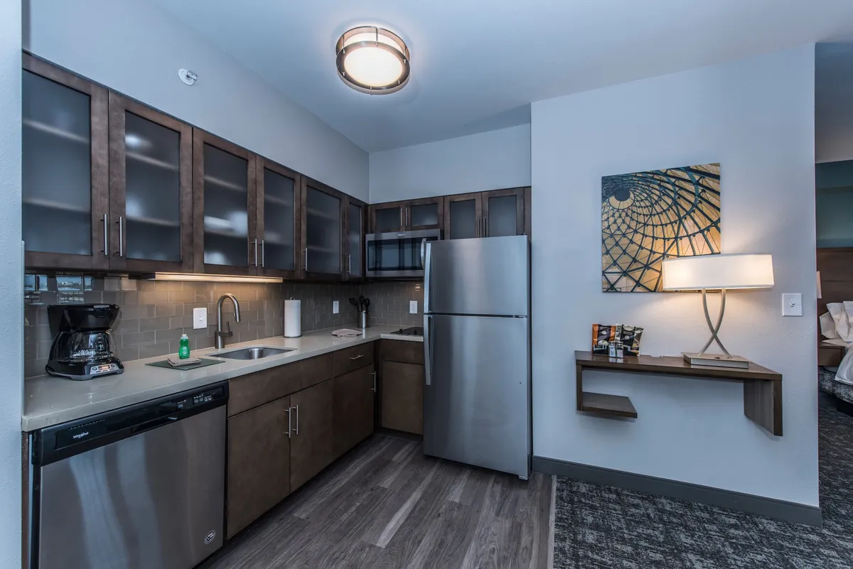 A cozy, contemporary kitchen scene with a stainless steel refrigerator, sleek wooden cabinetry, a prominent coffee maker, and warm lighting. The setup suggests a relaxed atmosphere for morning coffee or preparing light meals, with artwork adding a personal touch.