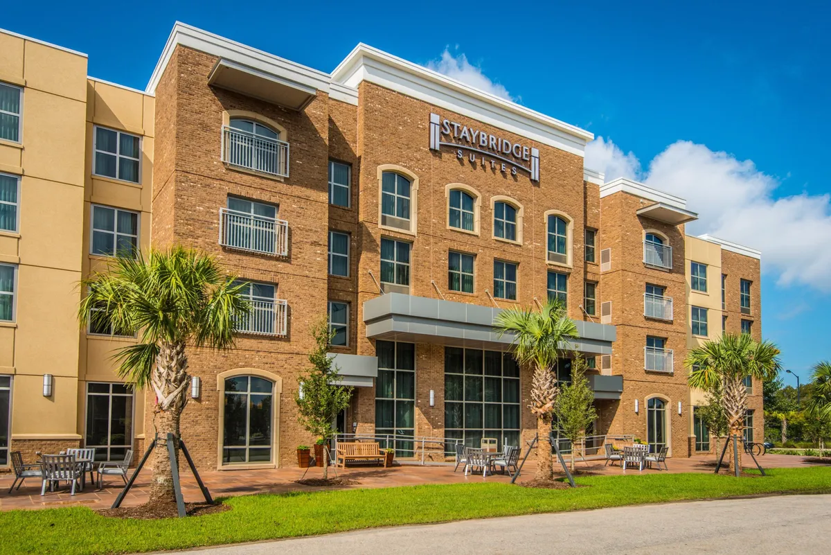 This image features the Staybridge Suites sign framed by lush palm trees, with an inviting outdoor seating area in the foreground. The scene exudes a welcoming and friendly atmosphere, giving a clear sense of arrival at the property and its connection to the outdoors.