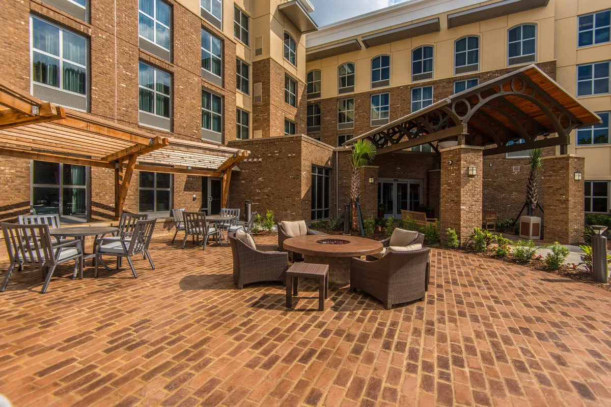 An exterior brick patio showcases a cozy fire pit surrounded by comfortable seating, all nestled beneath an overhead structure. The space is designed for social gatherings and relaxation, with a warm, communal mood enhanced by the firelight.