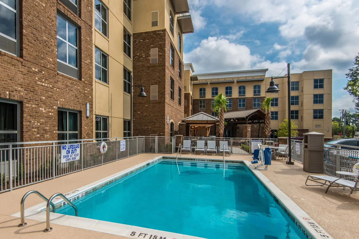 A vibrant outdoor swimming pool surrounded by lounge chairs and palm trees, with a poolside cabana providing shaded relaxation. The scene captures people enjoying the water, sunbathing, and socializing in a relaxed, resort-like setting under sunny skies.