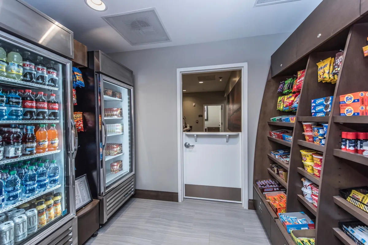 The image shows an interior snack shop with refrigerated drink coolers, well-stocked snack shelves, and a doorway leading to another part of the hotel. The setting is casual and relaxed, designed for quick grab-and-go purchases—ideal for busy guests on tight schedules.