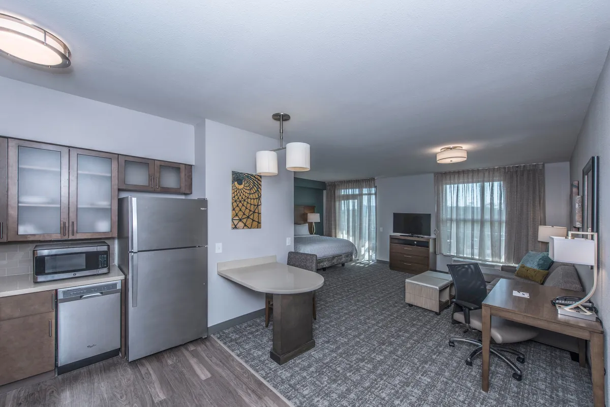 The image showcases a bright and welcoming studio interior, featuring a cozy bed, a compact kitchen area perfect for home-cooked meals, a dining table for two, a dedicated work desk, and a television—all arranged to maximize comfort and functionality in an inviting space.