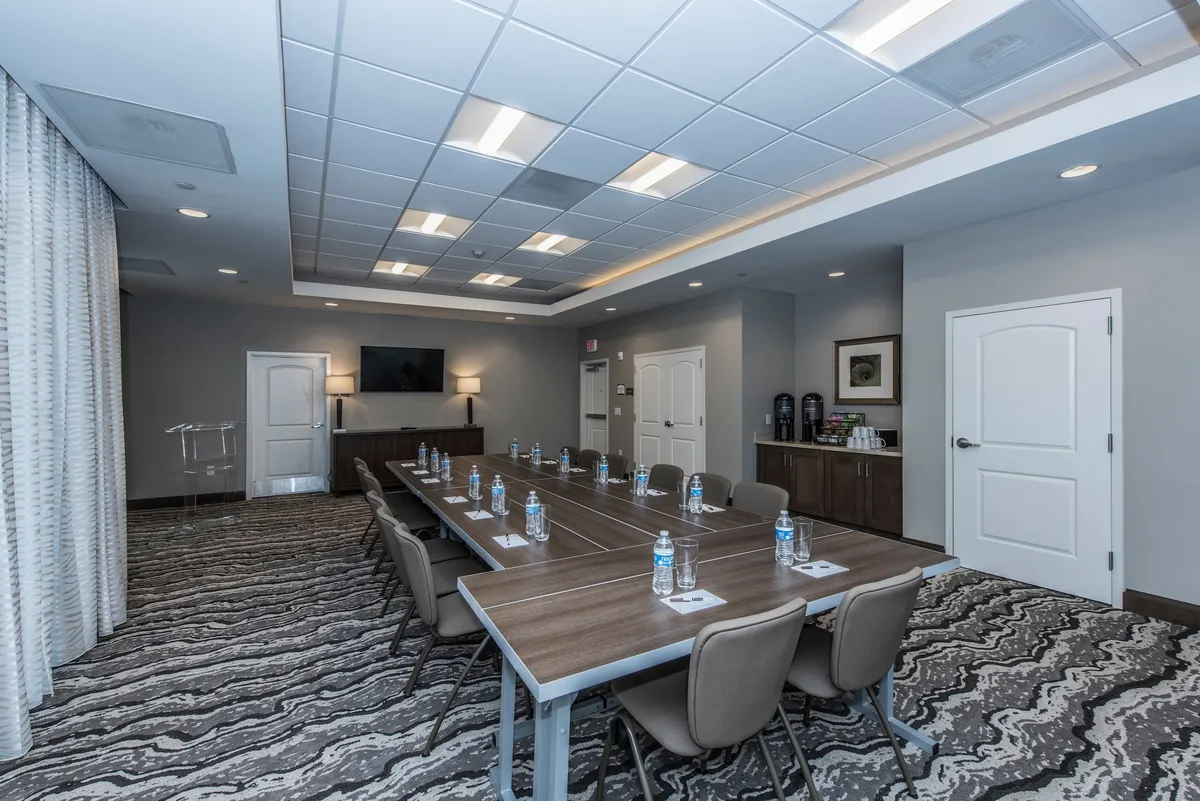 A modern business center featuring a long conference table, a television screen for presentations, and water bottles set out for attendees. The space is designed for collaboration, networking, and focused productivity.
